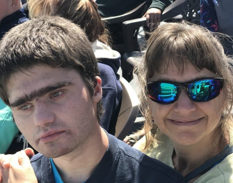 A mom hugs her young adult son. She’s wearing sunglasses and smiling.