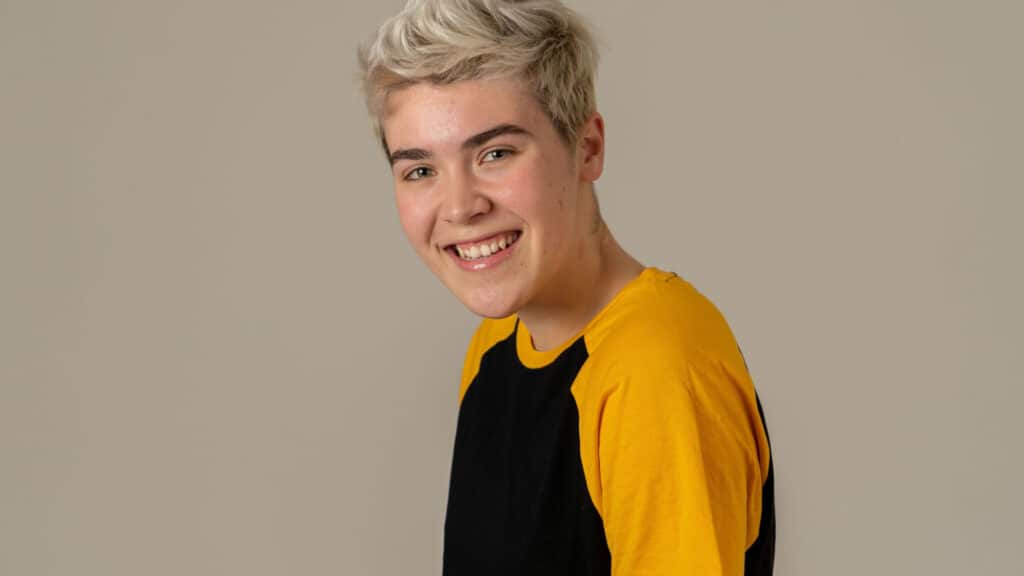 A white teenager, sideways to the camera, has their head turned and is smiling at the camera. They have short, spiky blonde hair.