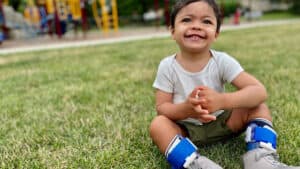 A young child with leg braces sits smiling in the grass