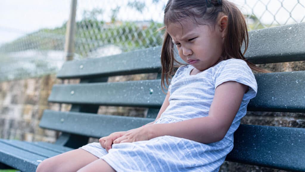 A small child with pigtails alone on a bench. She looks very unhappy.