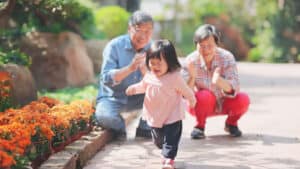 A toddler walks in a park while her grandparents cheer her on in the background. Walking and talking are examples of child development milestones.