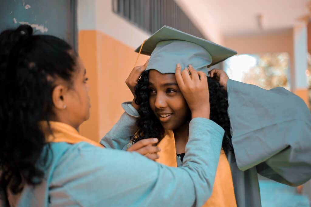 Young black woman gets ready for graduation. Her friend adjusts her cap.