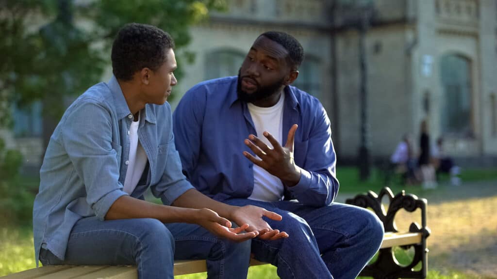 A young Black man sits on a bench in the sunshine listening to his dad. The men are leaning towards each other and are dressed casually.