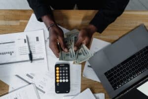 A person counts their money at a desk with a computer, calculator and bank statement nearby.
