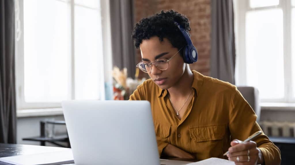 A young Black woman with headphones on is sitting at a table looking intently at her laptop. She is taking notes with pen and paper.
