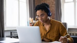 A young Black woman with headphones on is sitting at a table looking intently at her laptop. She is taking notes with pen and paper.