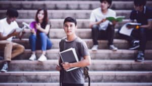 A young man with a backpack over one shoulder and books in his arms.