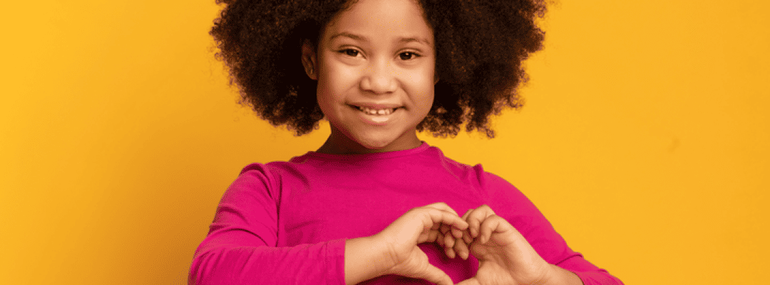 [Image Description: A black adolescent girl with brown, kinky, curly hair is smiling towards a camera in front of a bright, orange background. She's created a heart with her hands positioned on the left side of her chest. She's wearing a magenta lon…