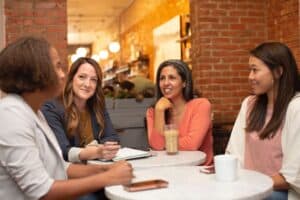 4 women are sitting at small round tables drinking coffee and talking.