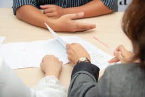 Three people sit at a desk with paperwork between them. Only their hands are visible.