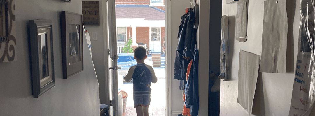 [Image Description: A pale skin adolescent boy is standing in the doorway with his back towards the camera. He's wearing a blue shirt with white long sleeves and grey shorts above the knees. In front of him is a house and beaming sunlight. Behind hi…