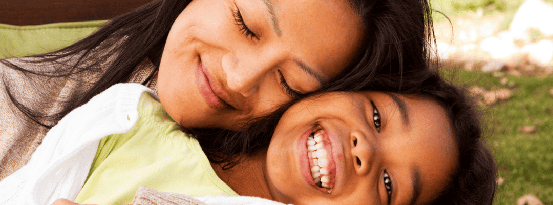 [Image Description: A light skin woman with fine, black hair holding a brown skin adolescent girl in her embrace. The woman's eyes are closed as she smiles with her head against the girl's cheek. The woman is wearing an oatmeal color sweater. The gi…