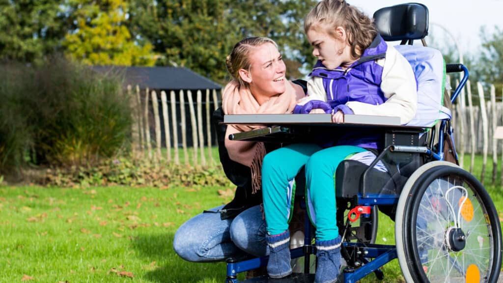 A girl in a wheelchair leans over to listen to a woman kneeling down next to her. They are outside on the grass.