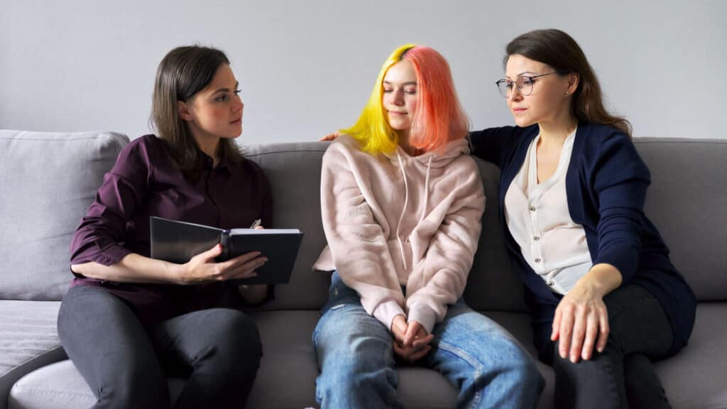 A worker from a social service agency for adults with disabilities is showing a book to a girl with disabilities and her mom as they sit on the couch.