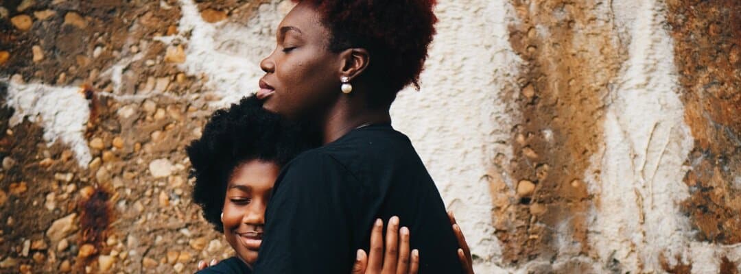 [Image Description: A Black woman and a Black girl are hugging with their eyes closed. The girl is smiling and has her head placed on the woman’s chest. She’s a dark blue shirt and her hair’s a dark brown afro. The woman has a slight smile. She has …