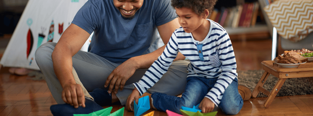 Image shows a Black floortime [capitalize?] therapist smiling, seated crossed legged playing with paper boats on the floor with a 6 year old boy. The therapist has black twists and is wearing a blue shirt and grey pants with black ankle socks. The b…