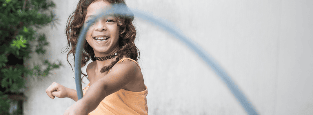 [Image Description: A light skin girl is smiling while holding a grey hula hoop on her wrist. She has brown shoulder length curly hair. She’s wearing a black choker with an orange tank top and hot pink bottoms. Behind her is a taupe colored wall and…