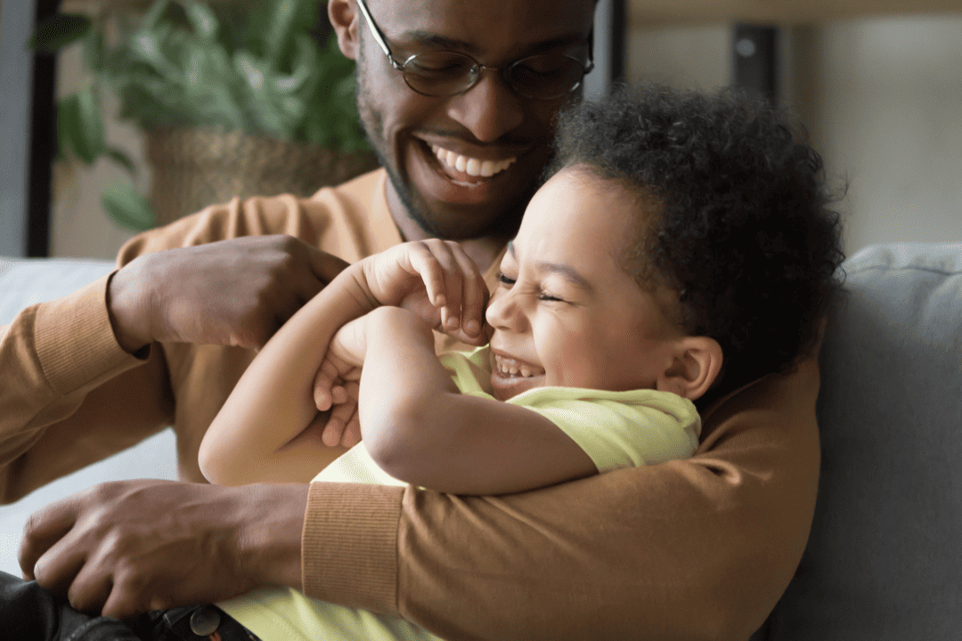 A son lays in his fathers arms as his father tickles him. They are both laughing.