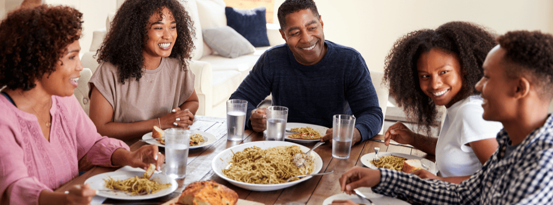 [Image Description: There's a Black family smiling and eating at a large, rectangular wooden table. In the center of the table is a white plate with spaghetti and to the left of the spaghetti is bread on a cutting board. There are 3 presenting women…