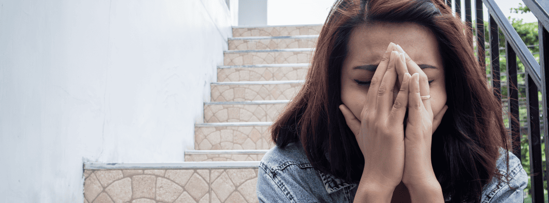 [Image Description: A light skin woman with auburn hair sitting at the bottom of a stone staircase outdoors beside a white wall. She's wearing a denim jacket. The woman is in distress. She's wincing and covering her face with both her arms near the …