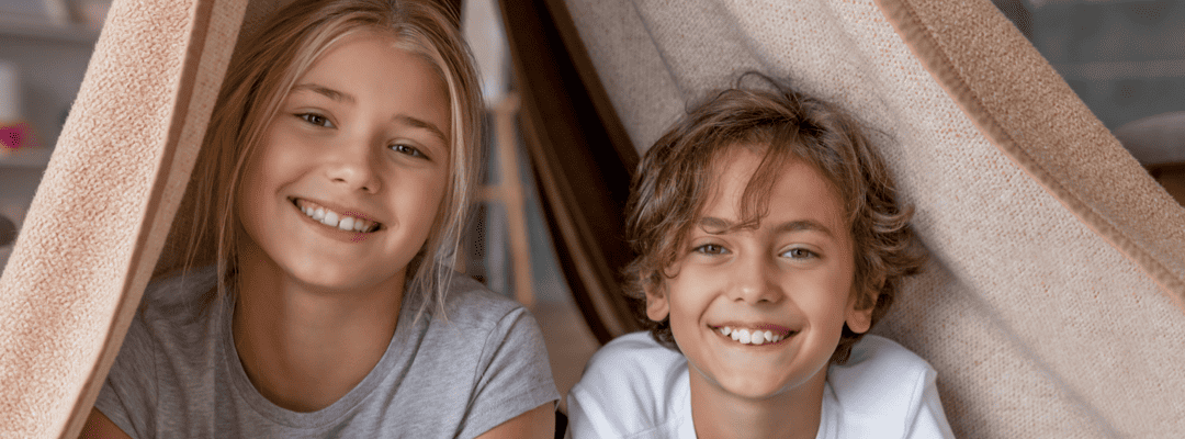[Image Description: A white 10 year old girl and a white 7 year old boy are lying in a blanket fort smiling towards the camera. The girl has straight, blonde hair and is wearing a grey shirt. The boy has sandy, blonde curly hair and is wearing a whi…
