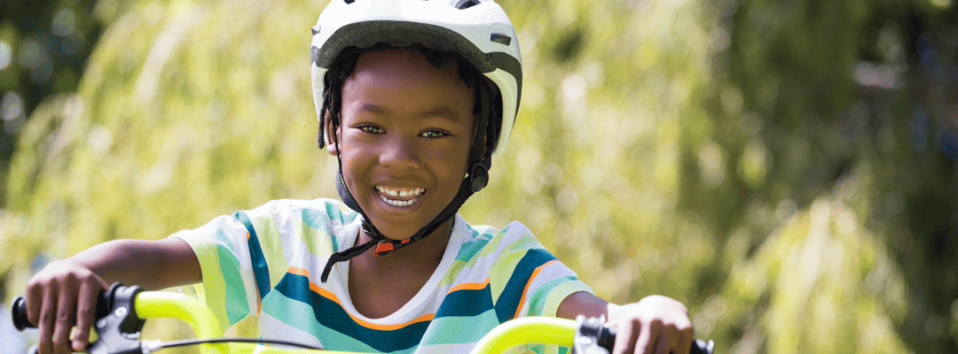 [Image Description: A Black presenting boy smiling holding his handlebars while riding a lime green bike with a white helmet. He’s wearing a green, teal, white, and orange striped shirt with red shorts cut off by the framing of the photograph. The b…