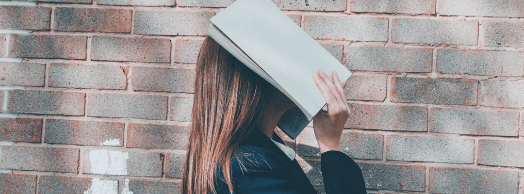 [Image Description: A pale skin girl with straight, brown, mid back length hair is standing with her head tilted upwards using her hands to hold a white book to cover her entire face. She’s wearing a navy blue jacket. She’s standing in front of a fa…