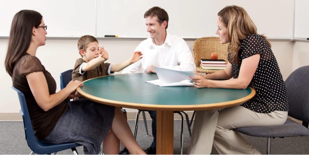 Parents, a teacher, and a small child sit together around a table in a classroom.