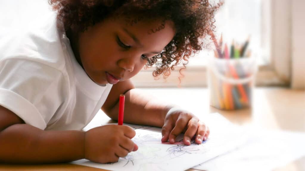 A preschooler in a white shirt leans over a piece of paper. She is drawing with a colored pencil.