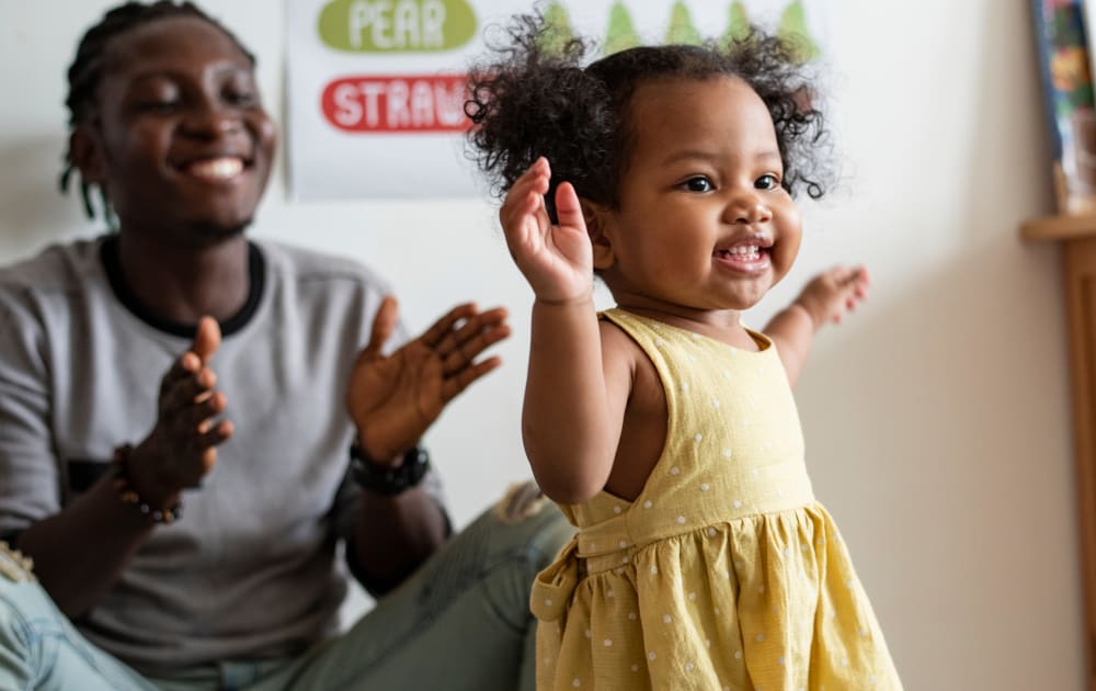 A toddler with pigtails holds her arms in the air smiling while her dad claps for her.