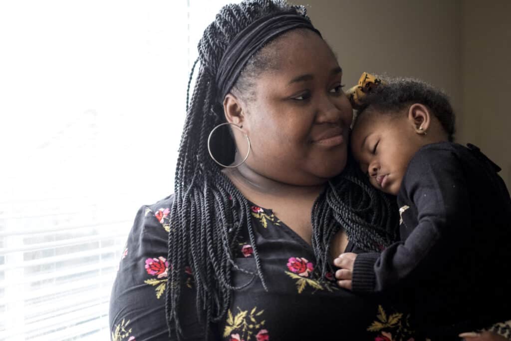 A Black woman with long braids holds her toddler, who is resting on her shoulder.