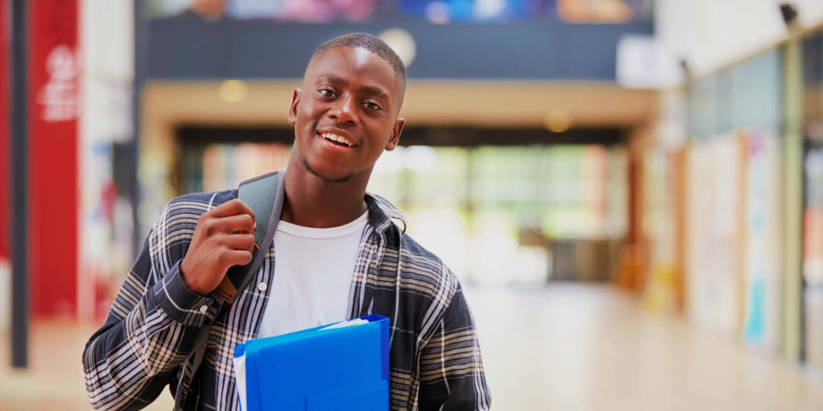 A young man with a backpack on is holding books and looking at the camera. He is in a school hallway.