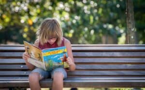 A child sits on a bench reading a book during the summer.