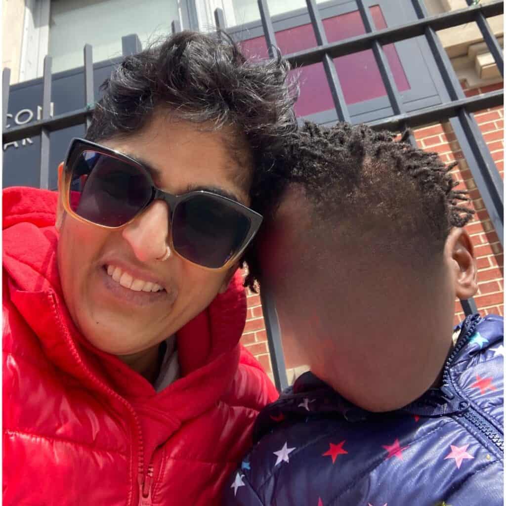 Shubha, a person of color smiling with their son, whose face is blurred for privacy reasons.
