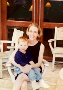 A white woman in a black dress holds a white toddler on her lap. They are both smiling at the camera.