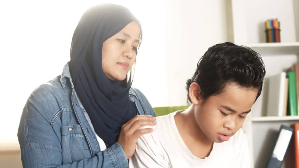 Una mujer en una camisa de jean y un hiyab consola a su hijo de edad escolar, quien se ve preocupado