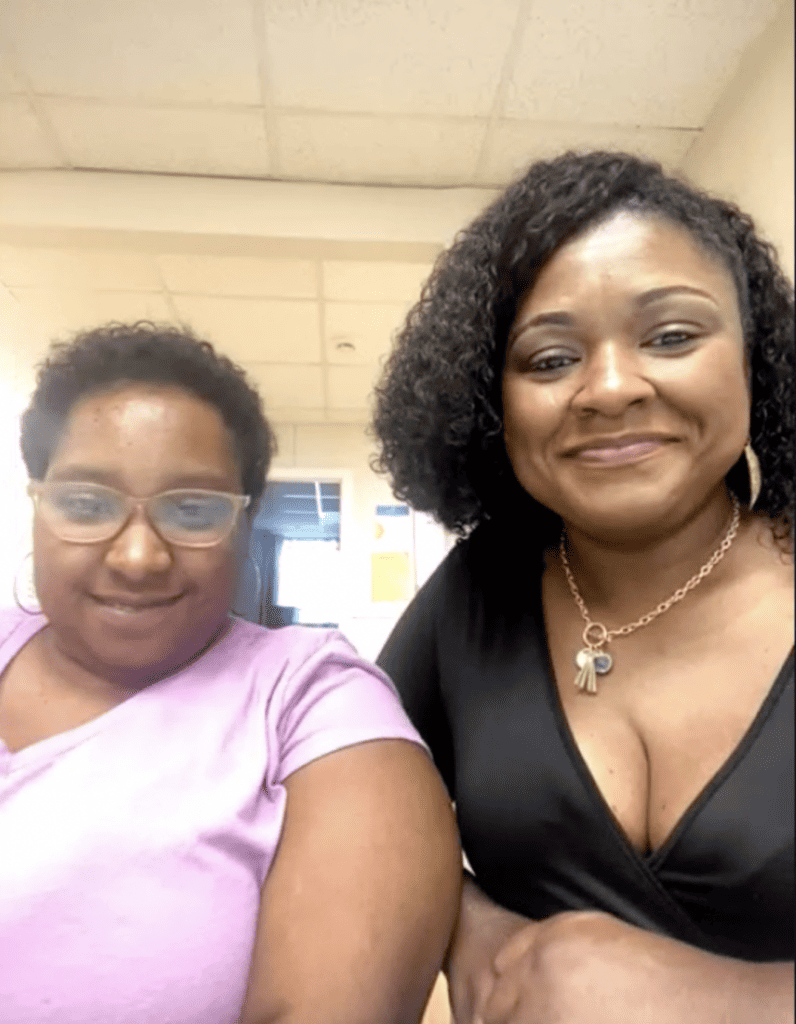 Black woman(mom) sitting next to her black daughter smiling.