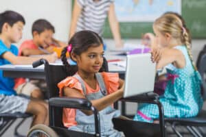 Una niña en una silla de ruedas usa una tableta mientras los otros alumnos escriben en sus mesas