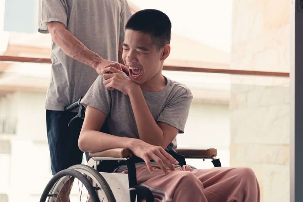 A teen in a wheelchair laughs and holds hands with his dad, who has his back
