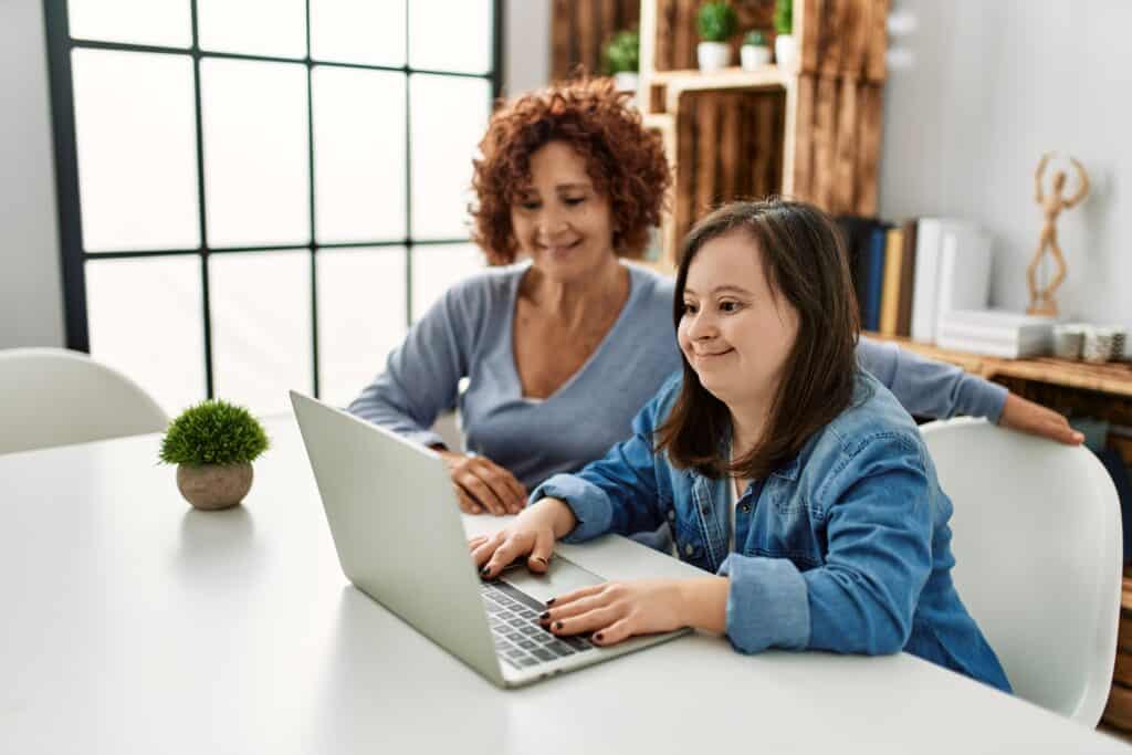 A girl with Down Syndrome smiles and works on a laptop with her mom