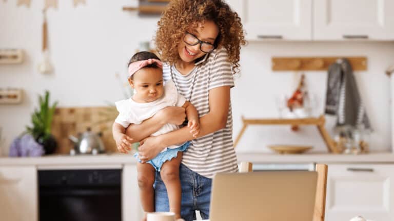 Woman holds baby while looking at her computer and talking on the phone.