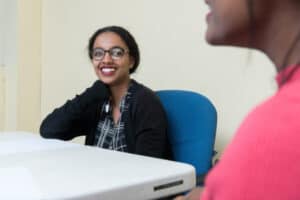 A teacher in glasses sitting at a conference table smiles at a parent seen in profile.
