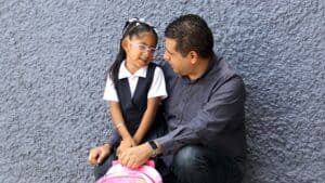 A dad has his arm around his little girl, who is holding a pink backpack