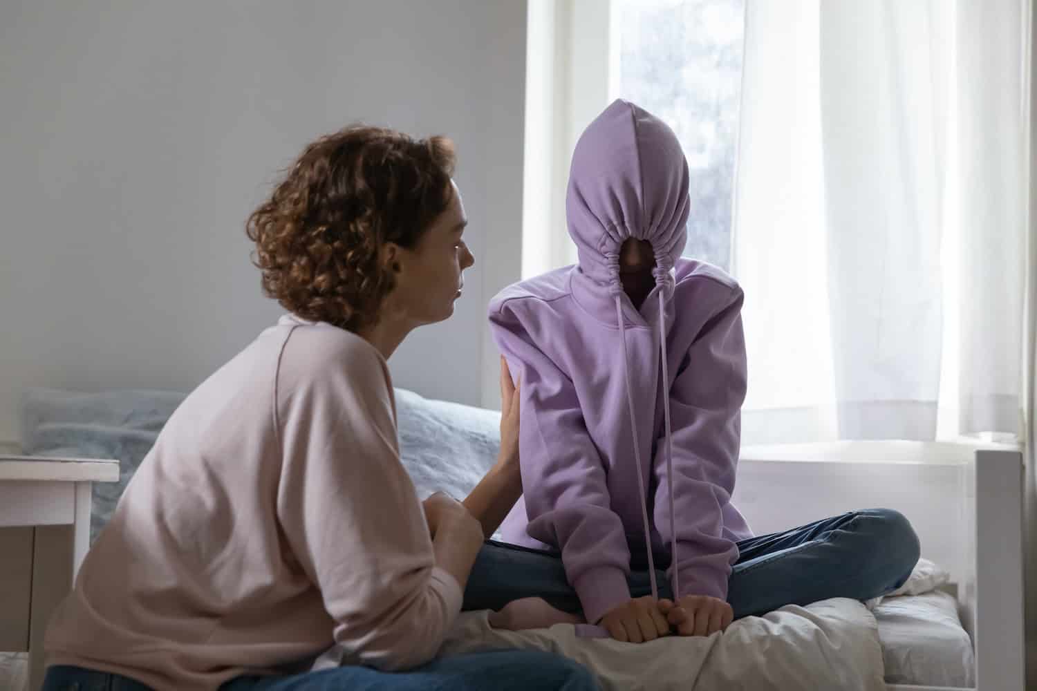 A girl sits on her bed with a hoodie over her face, refusing to go to school. Mom talks to her.