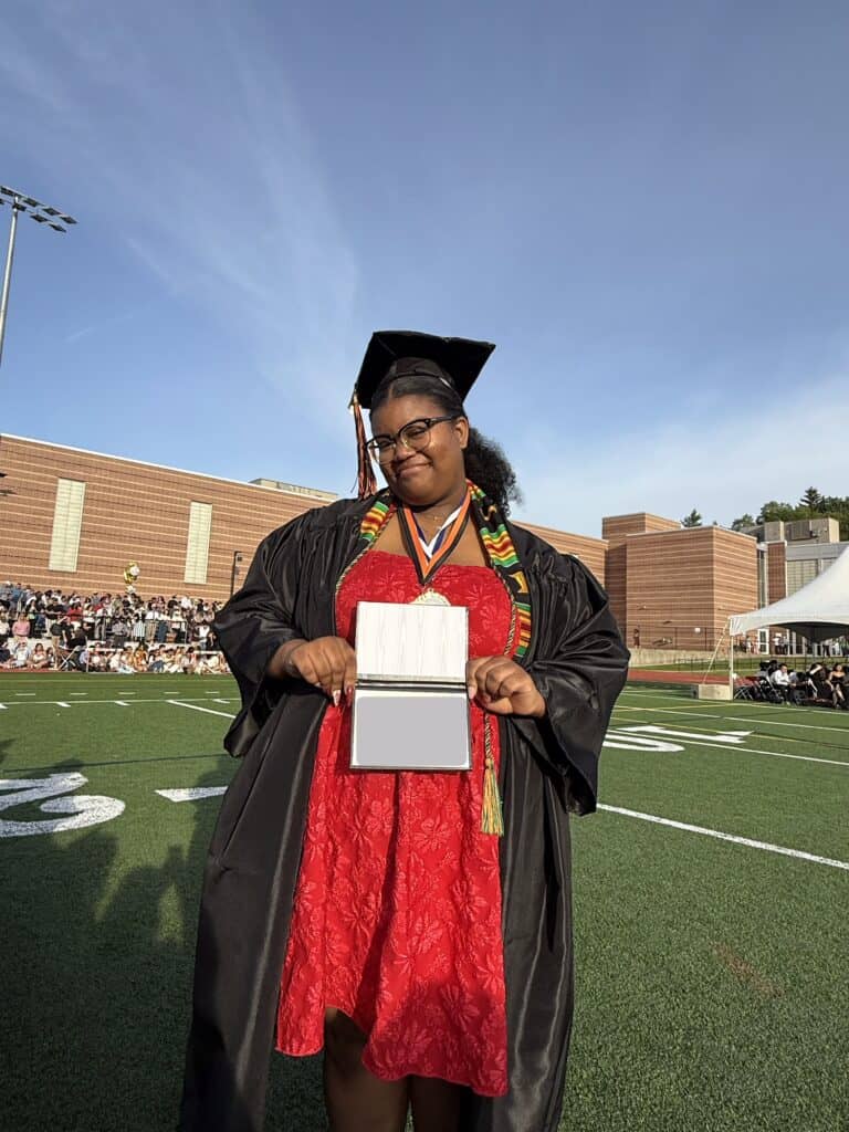 A smiling graduate in a red dress, cap and gown displays her diploma.