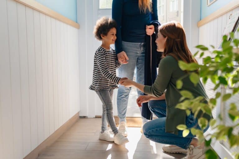 A young girl smiles as her mom introduces her to her new kindergarten teacher.